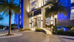 A modern hotel entrance with a curved driveway, palm trees, and a well-lit facade.