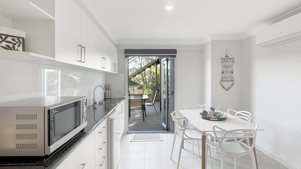 A modern kitchen with white cabinets, a built-in microwave, and a dining area with white chairs and a table. - Spacious Beach Studio With Alfresco Dining (Surf Beach)