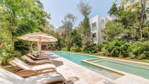 A poolside area with lounge chairs and a pergola umbrella, surrounded by lush greenery and a modern building in the background.