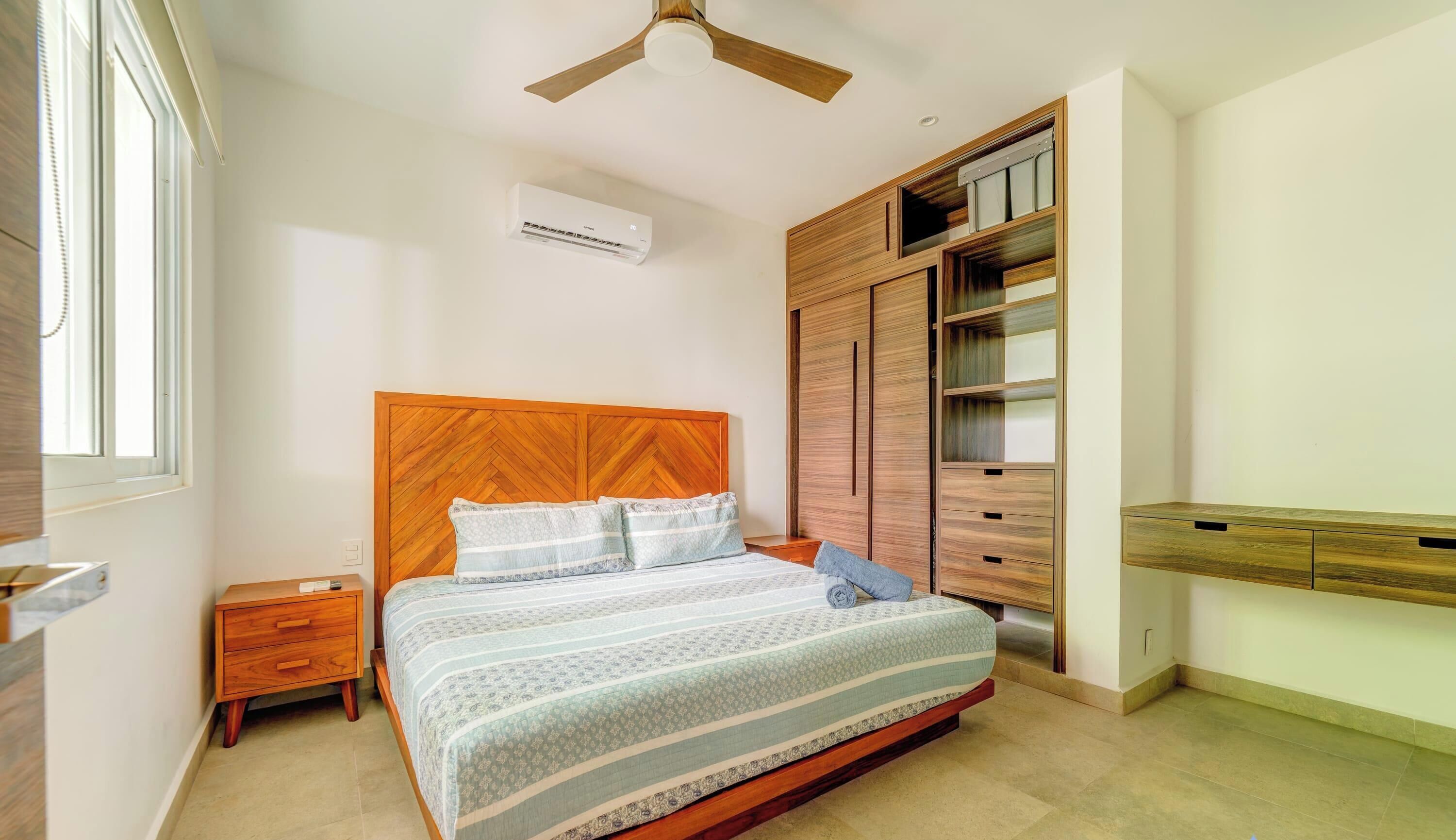 Quarto com cabeceira de madeira, cama com roupa de cama estampada, mesa de cabeceira, guarda-roupa com vários compartimentos e ventoinha no teto.