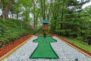 Un campo de mini golf con superficie de juego verde, una estructura de madera y una bandera roja que marca el hoyo.