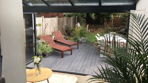 Uma estufa com deck de madeira, duas cadeiras de descanso, uma pequena mesa com um vaso de flores, e um jardim visível através do vidro.
