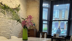 Uma área de refeições com mesa branca, bancos, um vaso verde com flores e uma janela com persianas.