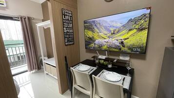 A dining area with a table set for two, a clock on the wall, and a scenic TV display.