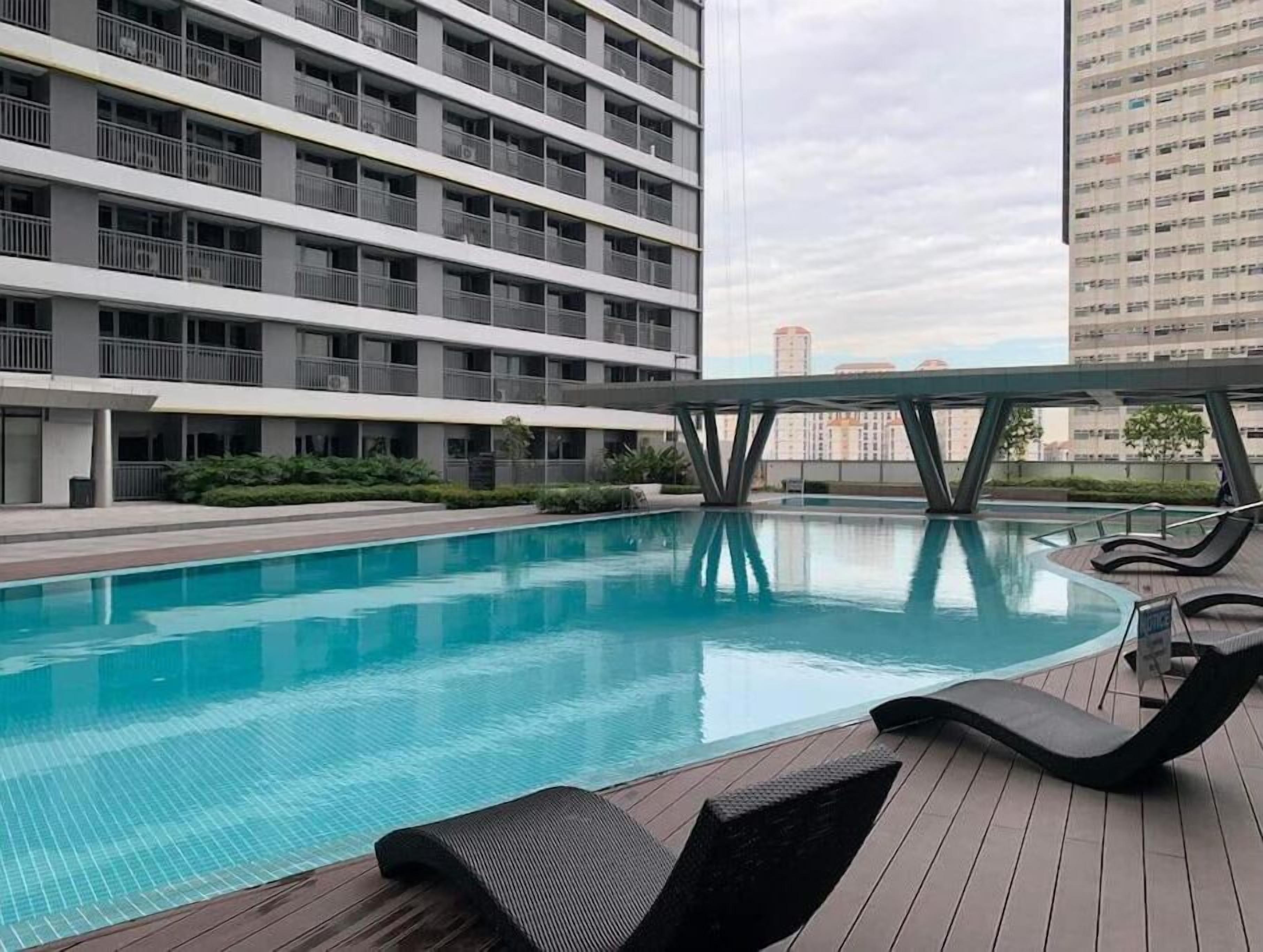 A swimming pool with a wooden deck and lounge chairs, surrounded by a multi-story building with balconies.