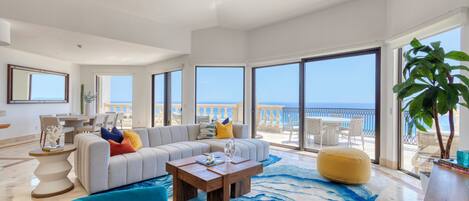 A spacious living room with a large sofa, a coffee table, and a view of the sea through large windows.
