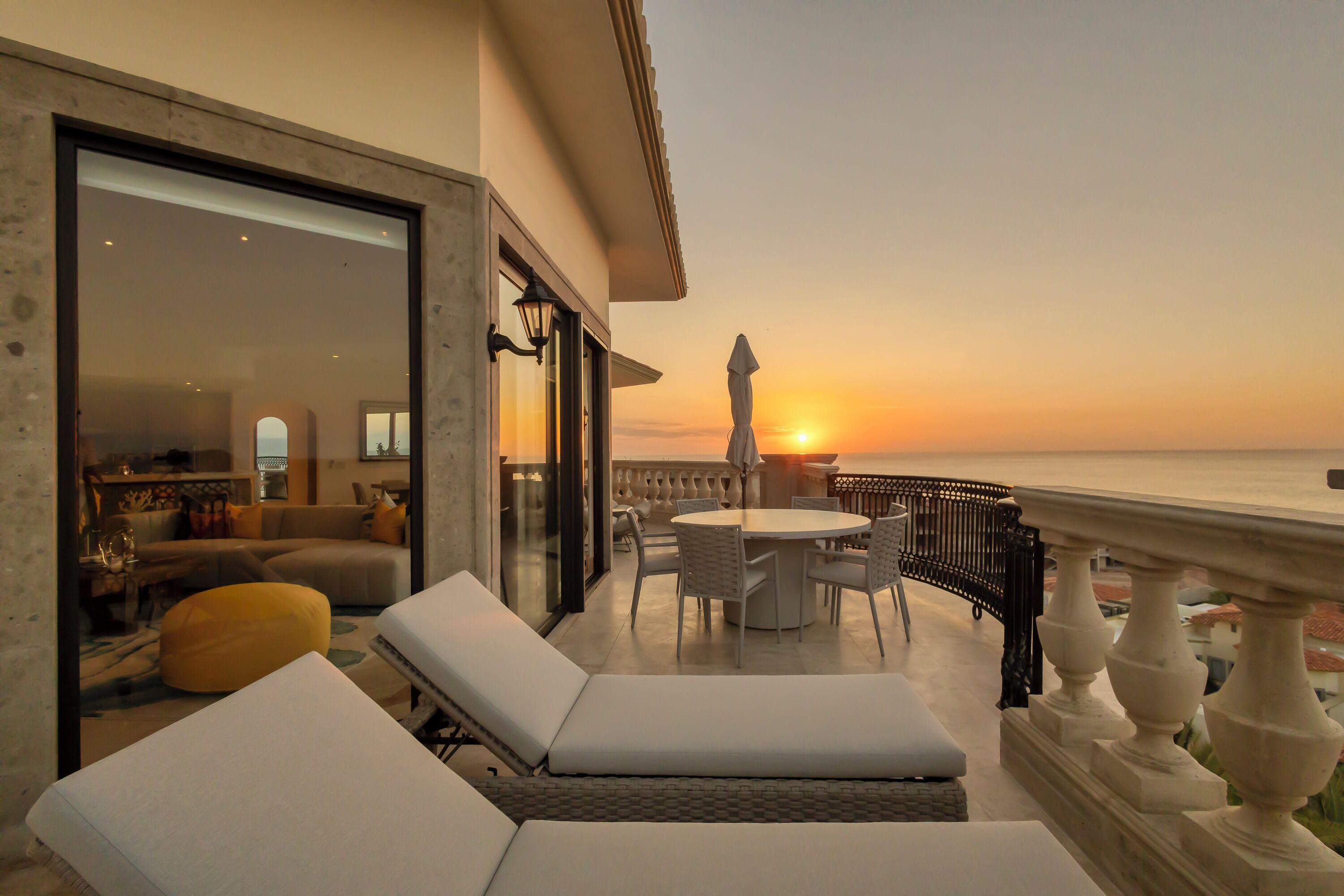 A balcony with lounge chairs, a table, and a view of the sunset over the sea.