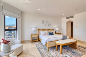 Un dormitorio con cama, mesitas de noche, una silla, un banco y vistas a edificios a través de una ventana.