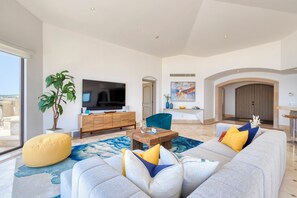 Una sala moderna con televisor de pantalla plana, un mueble de madera, un sofá con cojines de colores, una planta grande en maceta y vistas a través de un ventanal.