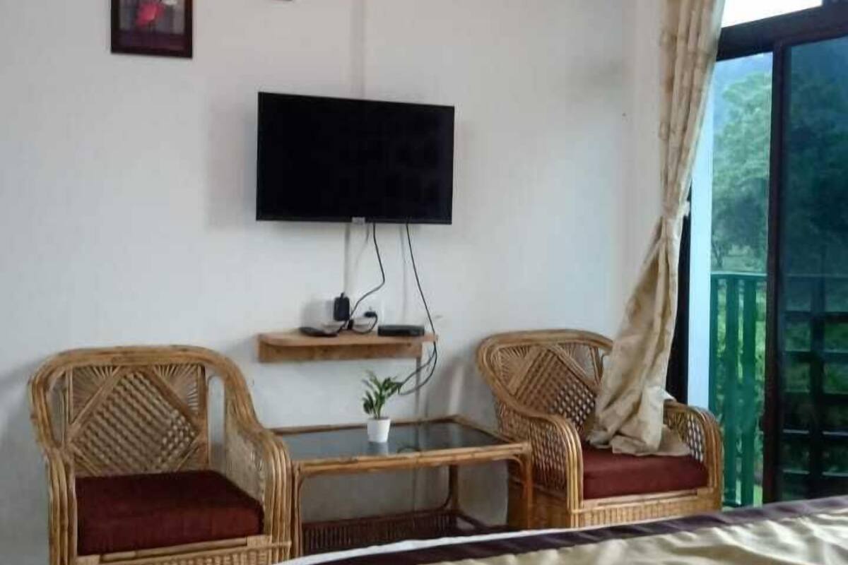 A bedroom with a bed, two wicker chairs, a small table, a TV, and a window with curtains.