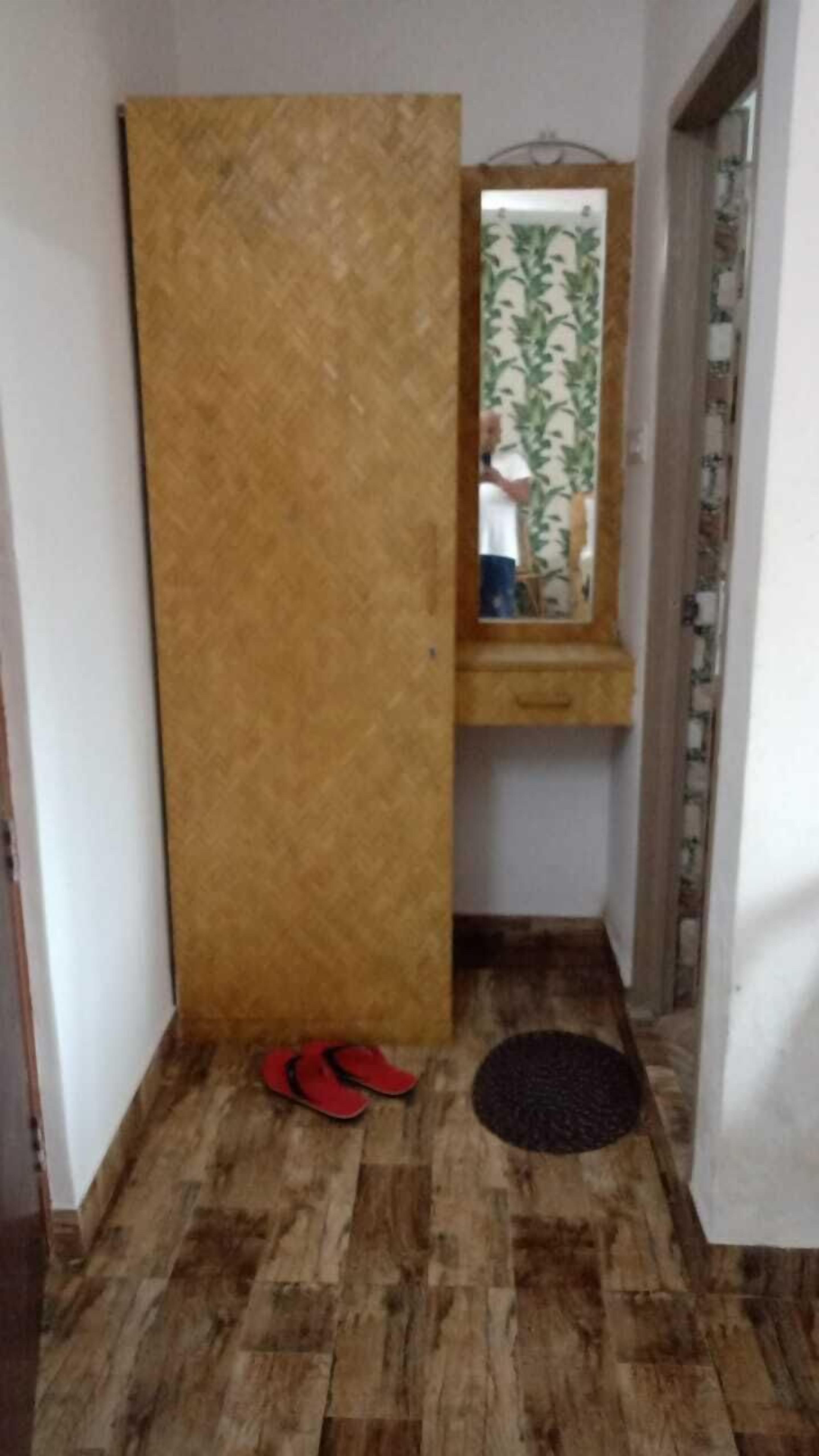 Une petite pièce avec une armoire en bois, un miroir, une petite étagère en bois, une paire de pantoufles rouges et un paillasson sur le sol.