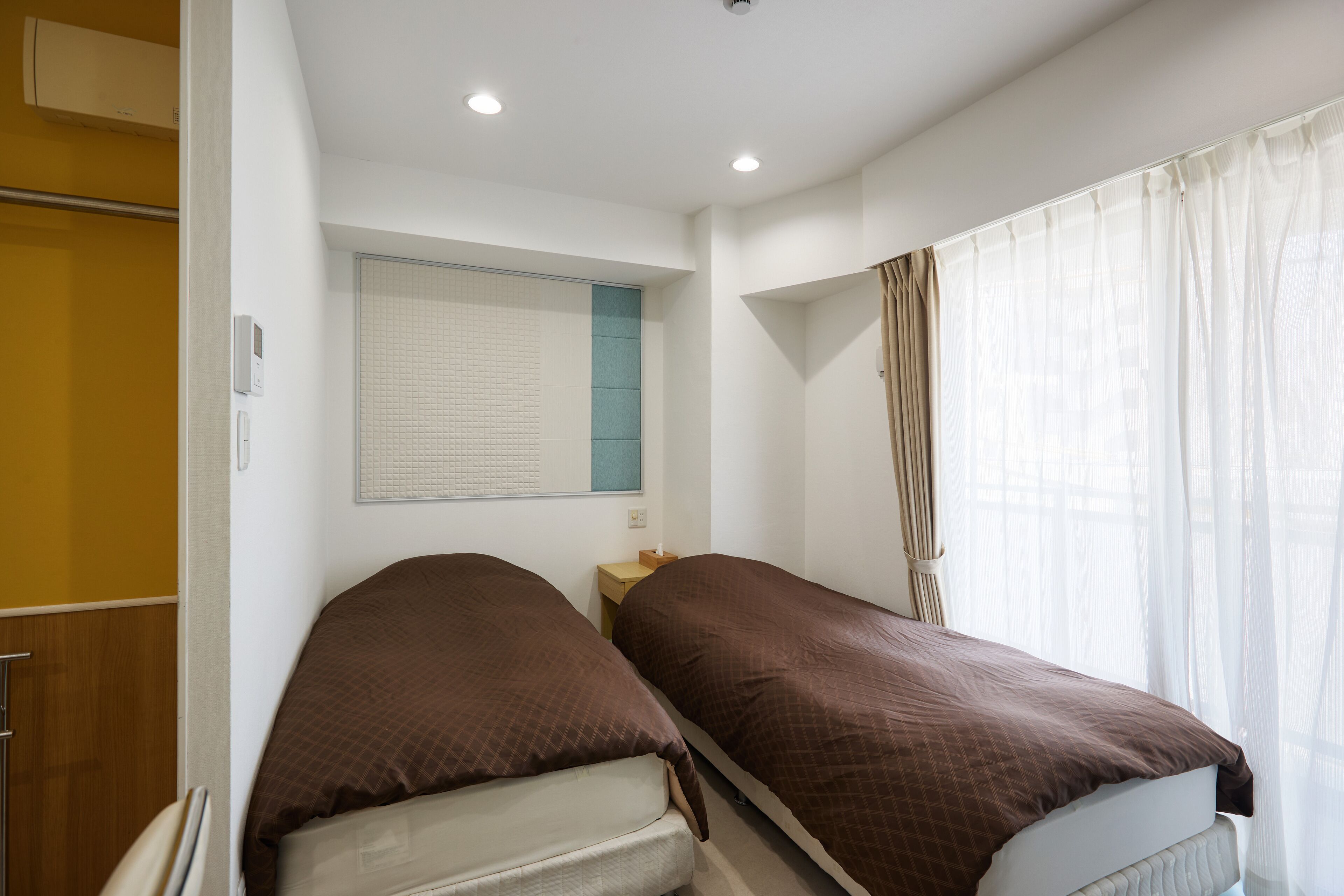 A hotel room with two single beds, a window with sheer curtains, a bedside table, and a wall-mounted air conditioner.