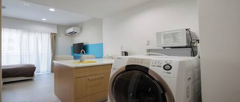 A compact hotel room with a washing machine, a bed, a small kitchenette, and a window with curtains.