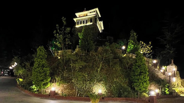 A well-lit outdoor area with a circular stone fountain, surrounded by trees and a building with illuminated windows. - The Jungle Story By Headquarter (Shimla)