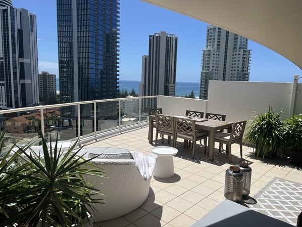 A balcony with a table and chairs, a potted plant, and a view of tall buildings and the ocean.