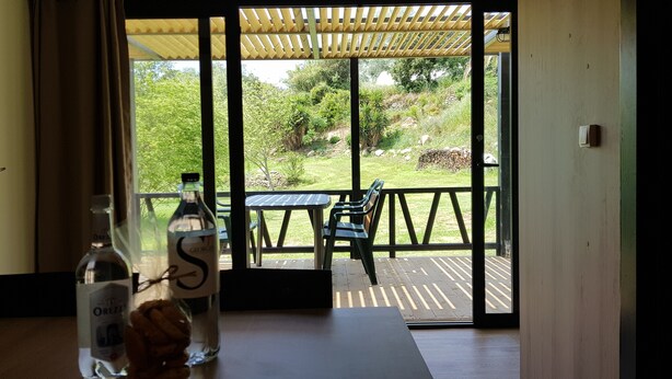 A room with a wooden table and chairs, a bottle of OREZZA, and a view of a garden through a glass door. - Logement Proche D'ajaccio et des Plages de Porticcio Pour 4 Personnes (Bastelicaccia)