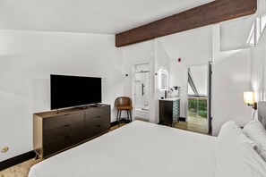 A modern bedroom with a large bed, a TV, a dresser, and a chair. There is a balcony with a view of the outdoors.
