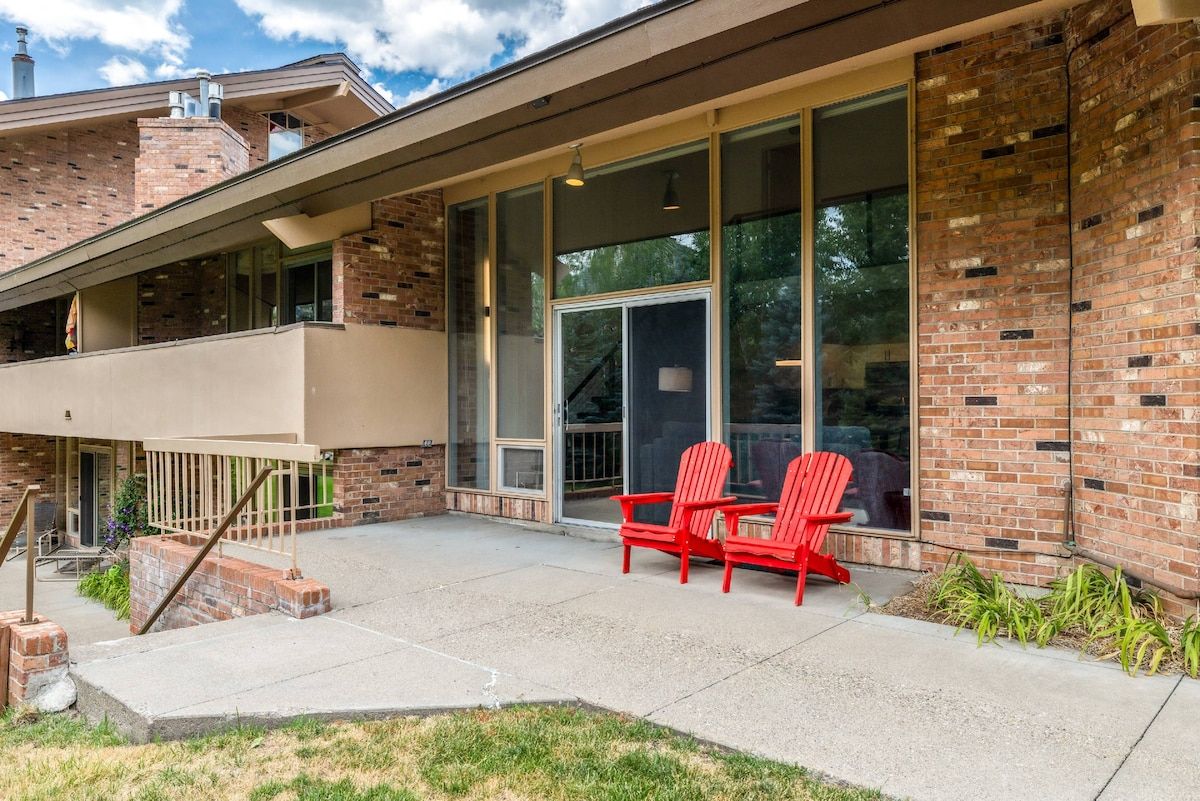 A modern building with a brick facade, large glass windows, and a covered patio area with red chairs.