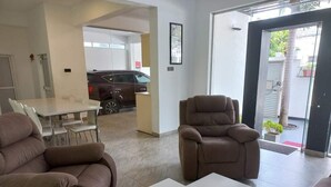 Een moderne woonkamer met een bank, fauteuil en salontafel. Er is een eethoek met stoelen en een tafel. Een auto staat buiten geparkeerd.