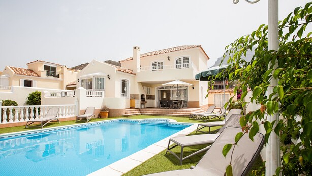 A swimming pool with lounge chairs, a patio area, and a large house with multiple balconies.