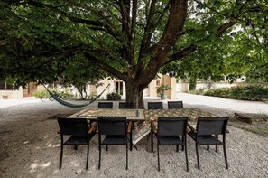 An outdoor seating area with a table and chairs under a large tree, surrounded by gravel and a building in the background.