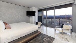 Una habitación de hotel moderna con una cama grande, un televisor de pantalla plana, un espacio de descanso con vistas a la ciudad y un balcón con panorama urbano.