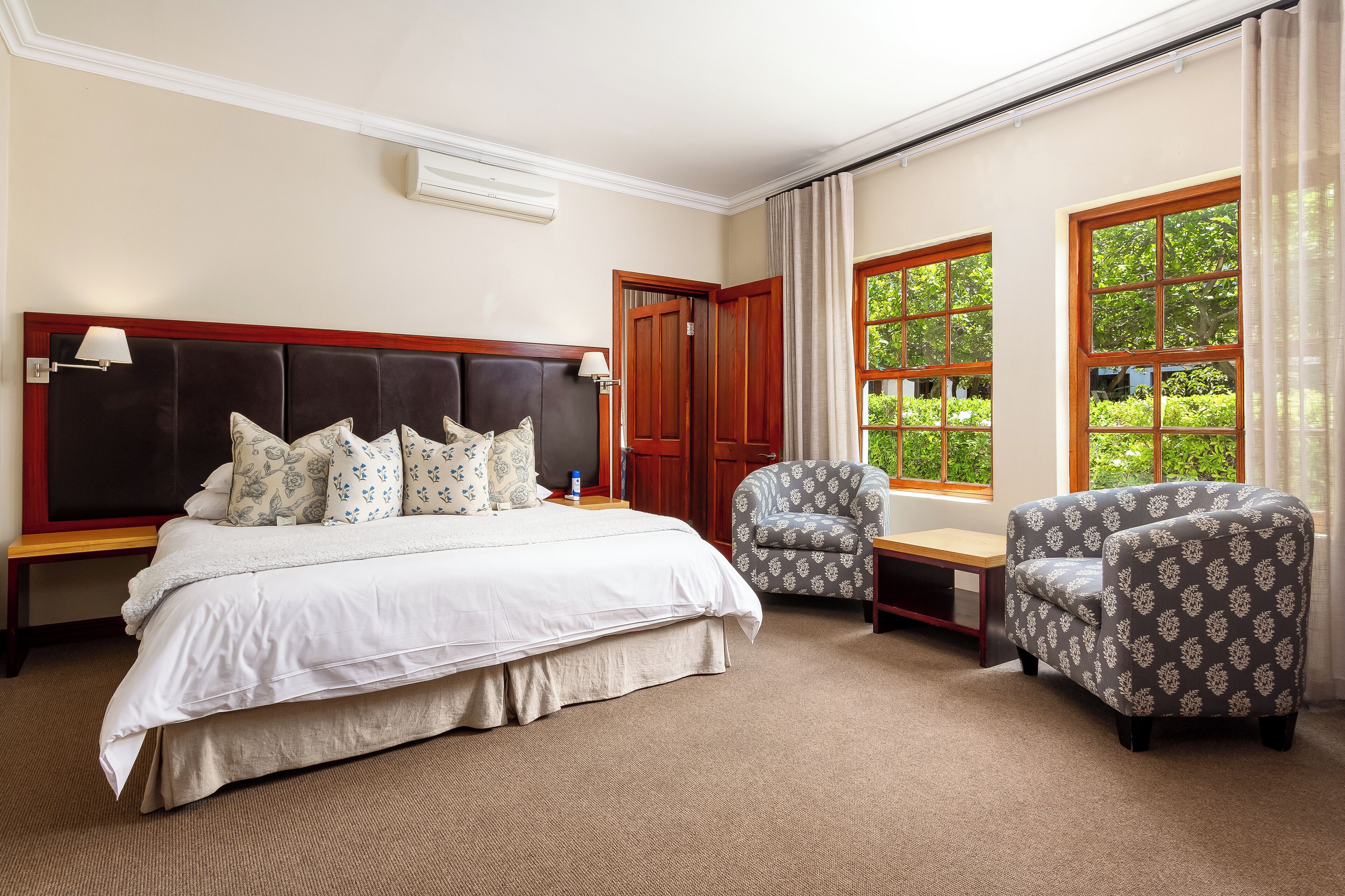 A hotel room with a large bed, two armchairs, a wooden nightstand, a bench, and a window with a view of greenery.