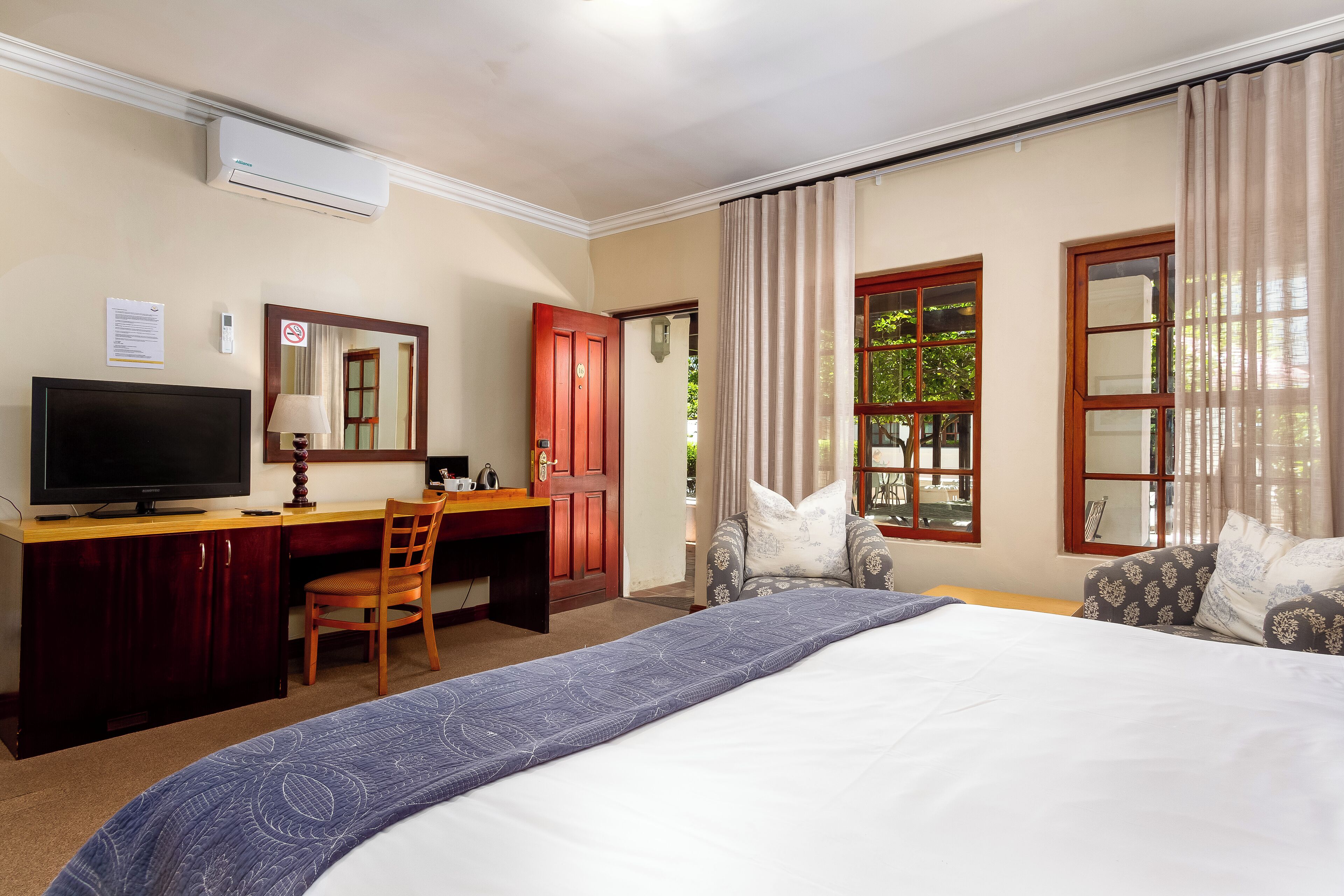 A hotel room with a bed, a desk with a television, a chair, a desk lamp, a window with curtains, and a door.