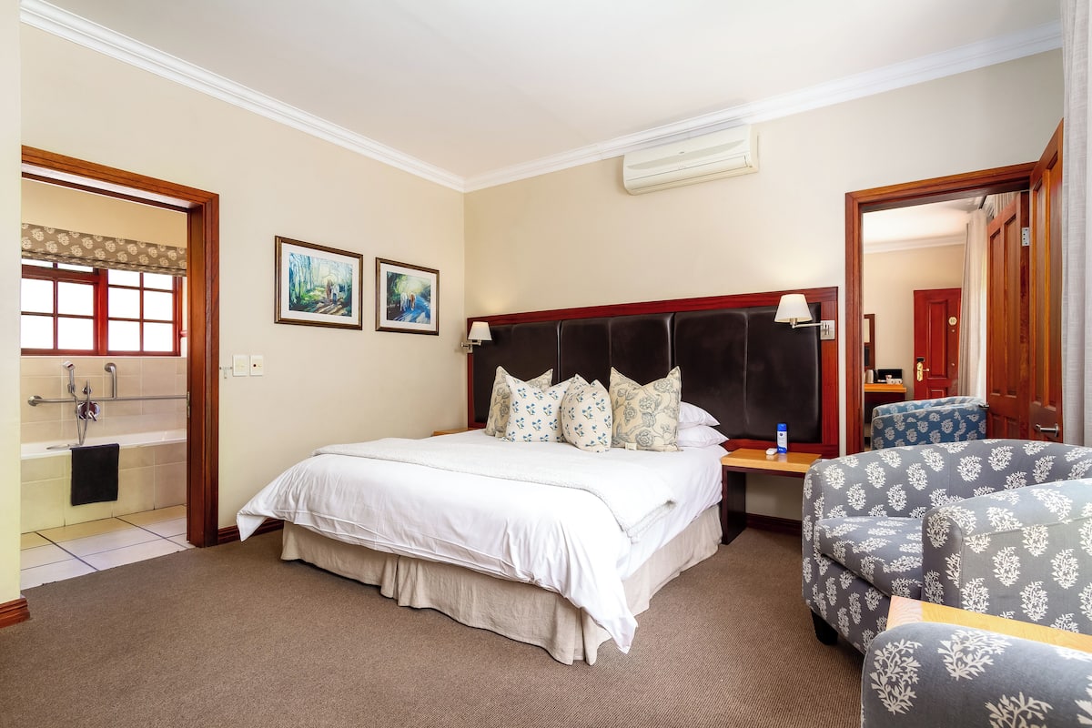 Habitación de hotel con una cama grande, una mesita de noche, una silla, un ventanal con cortinas y un cuadro en la pared.