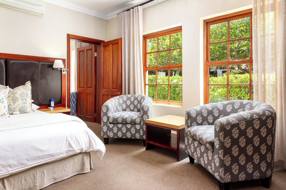 Habitación de hotel con una cama grande, dos sillones, una mesita y ventanas amplias con vistas a zonas verdes.