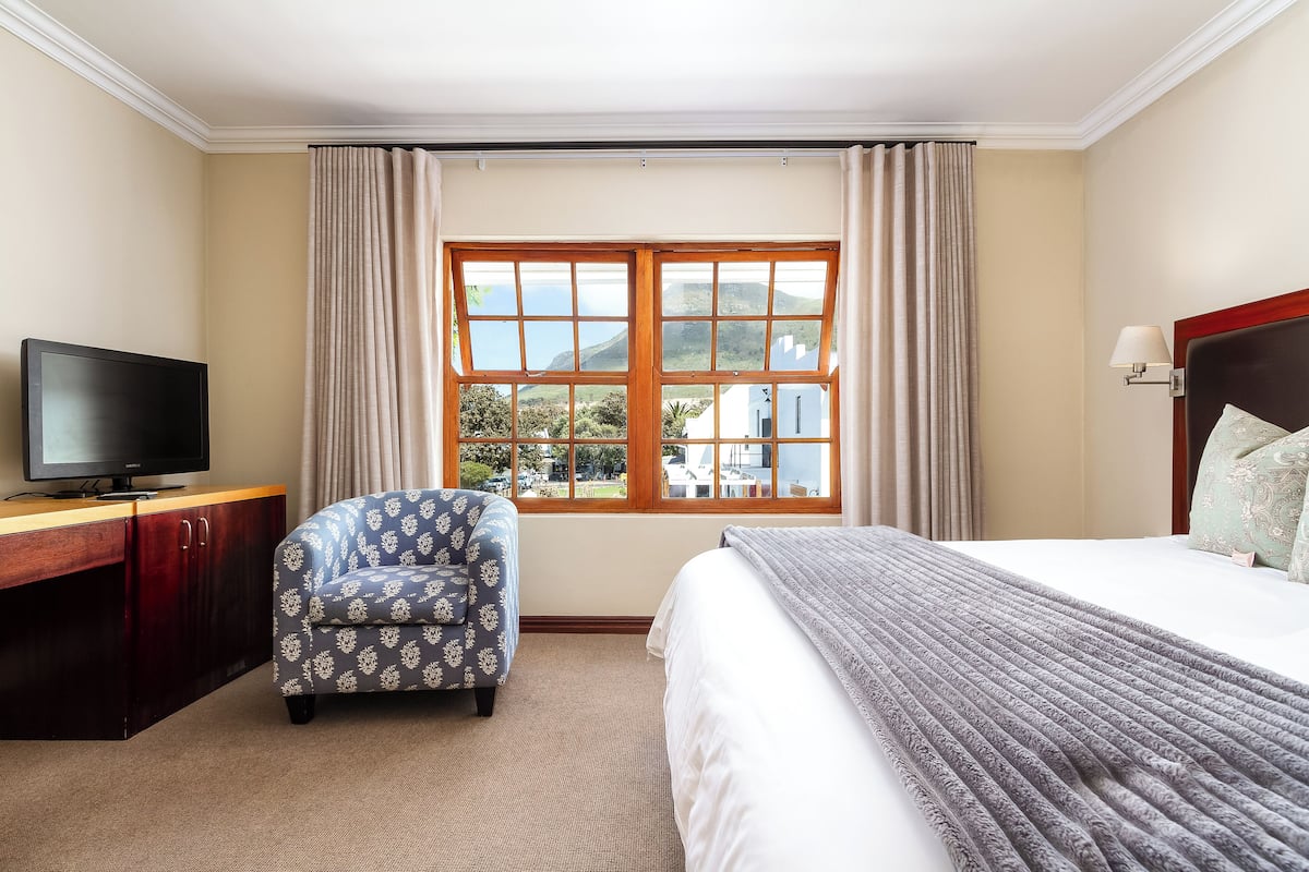Una habitación de hotel con una cama, un televisor sobre un escritorio de madera, un sillón estampado y un ventanal con cortinas.