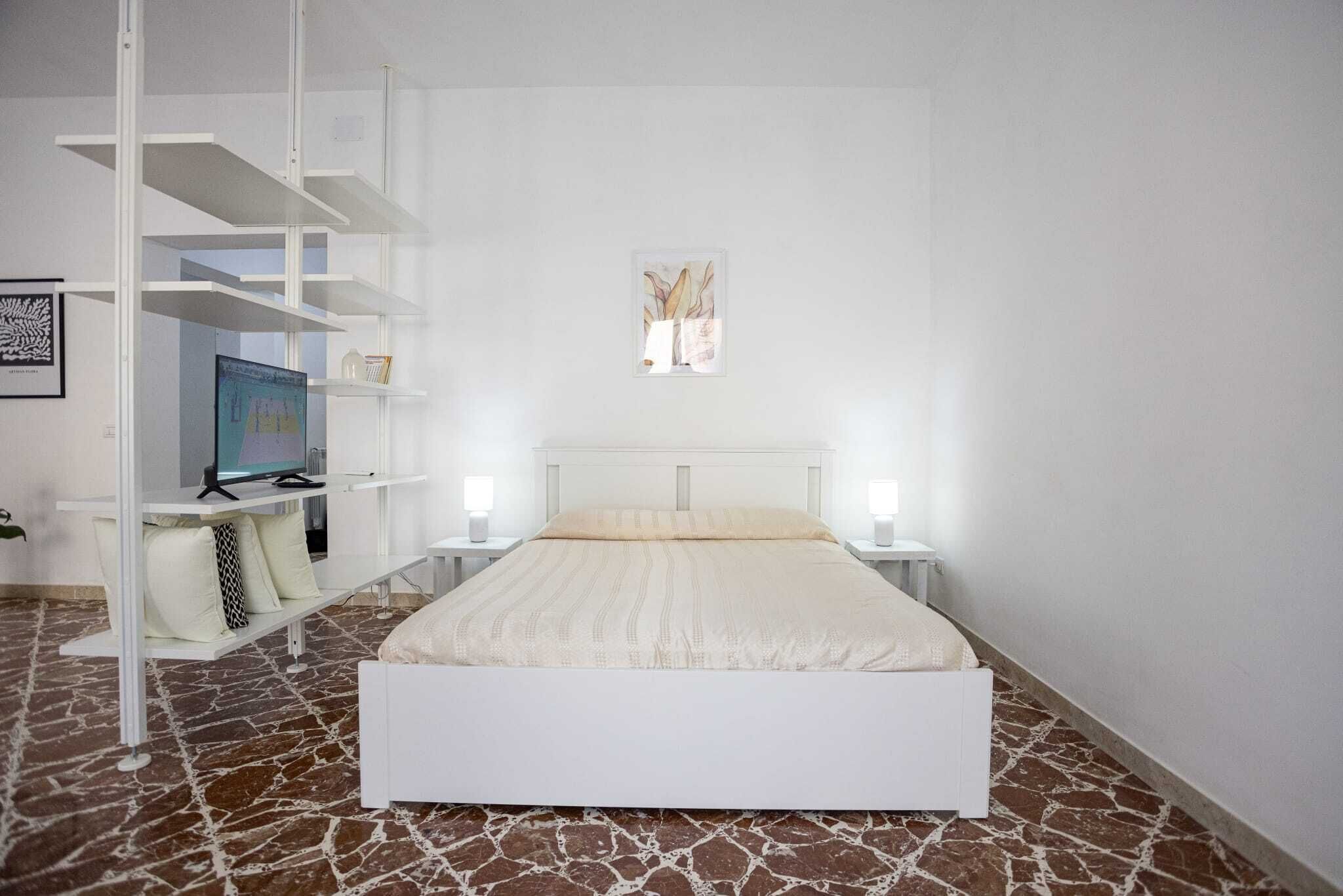 Ein Schlafzimmer mit einem weißen Bett, einem Schreibtisch mit Computer und einem Bücherregal.