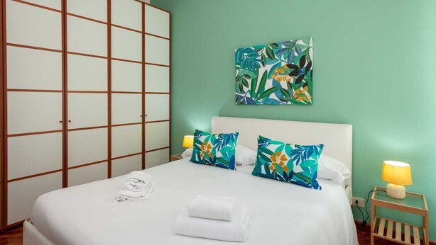 Un lit bien fait, avec du linge de lit blanc, deux coussins décoratifs à motifs tropicaux, une lampe de chevet et une œuvre d’art accrochée au mur au-dessus du lit.