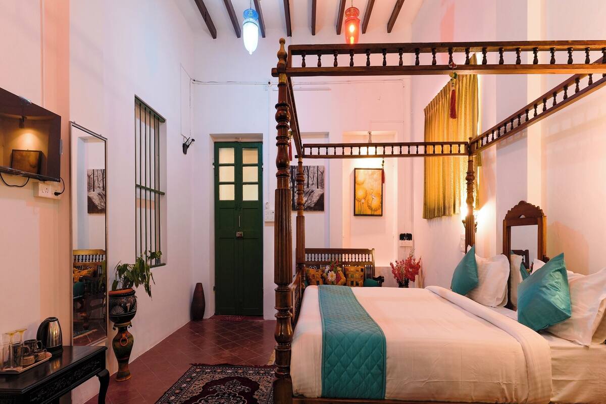 A traditional four-poster bed with a teal quilt, a wooden floor, a green door, and a mirror on the wall.