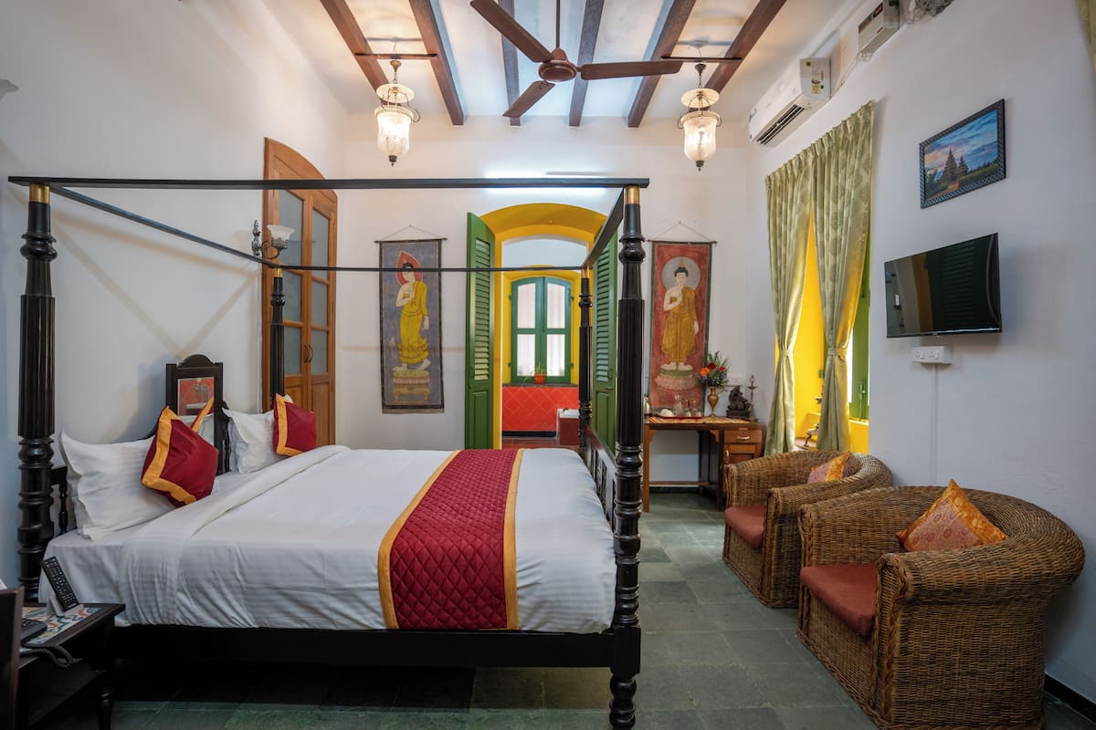 A four-poster bed with a red and white bedspread, a wooden headboard, and two wicker armchairs with red cushions.