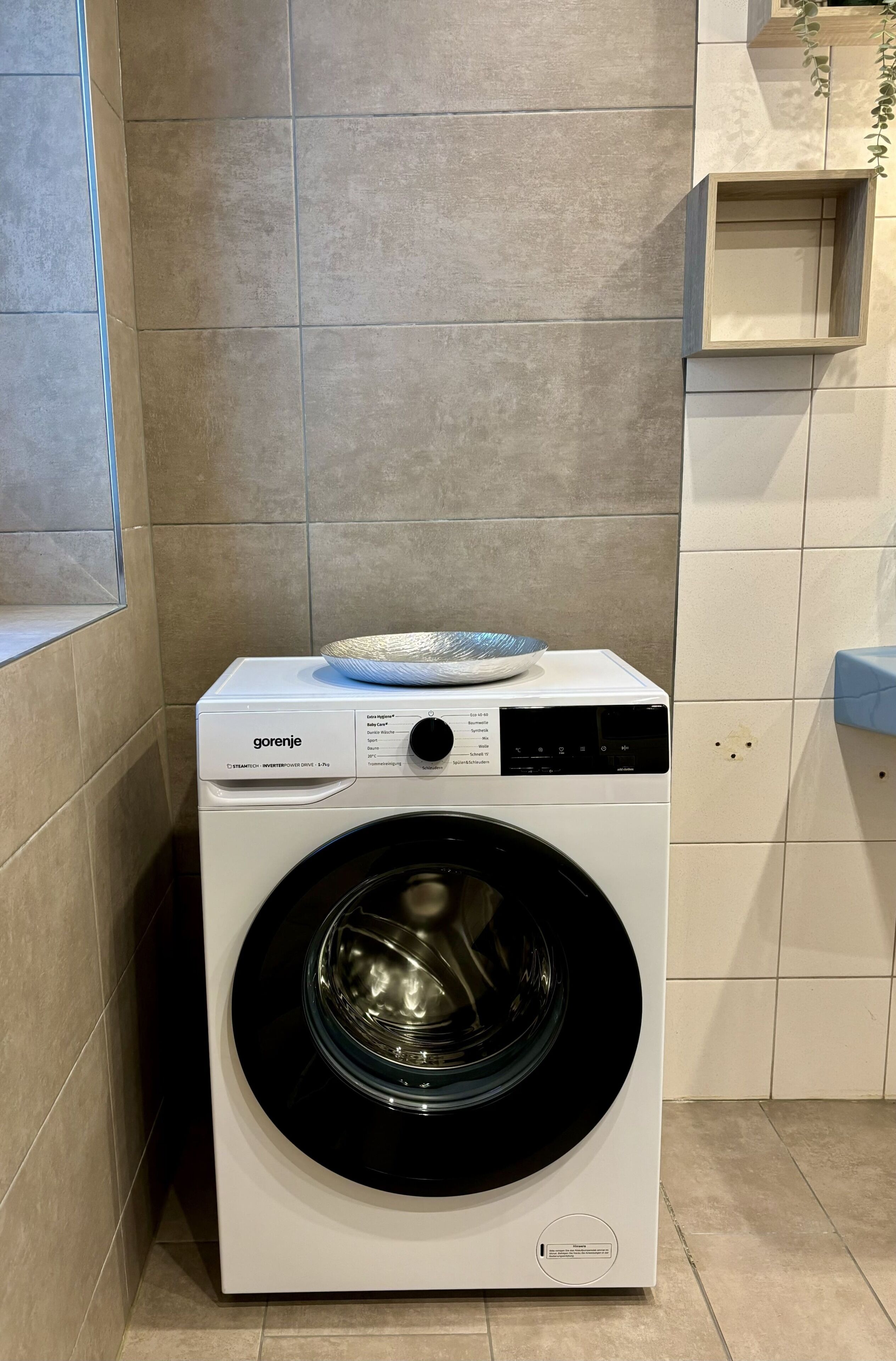 A front-loading washing machine with a control panel and a bowl on top, placed in a corner of a room with tiled walls and a window.