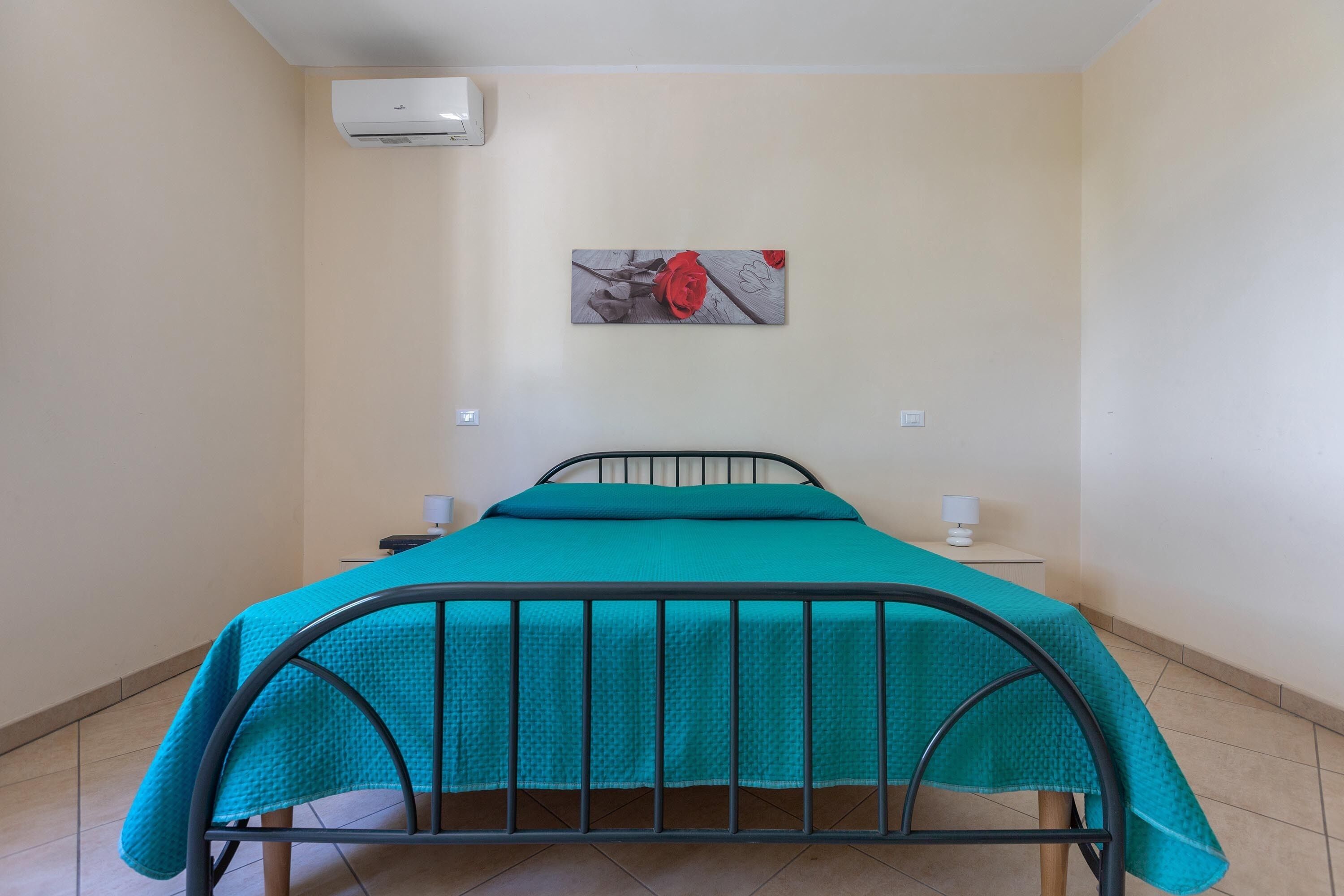 Ein Einzelbett mit türkisblau gestreuter Bettdecke, eine Wandklimaanlage und ein Gemälde einer roten Rose.