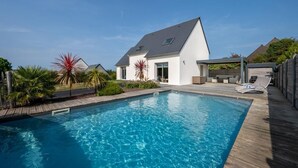 Una casa moderna con piscina, un porche cubierto y un jardín con plantas tropicales.