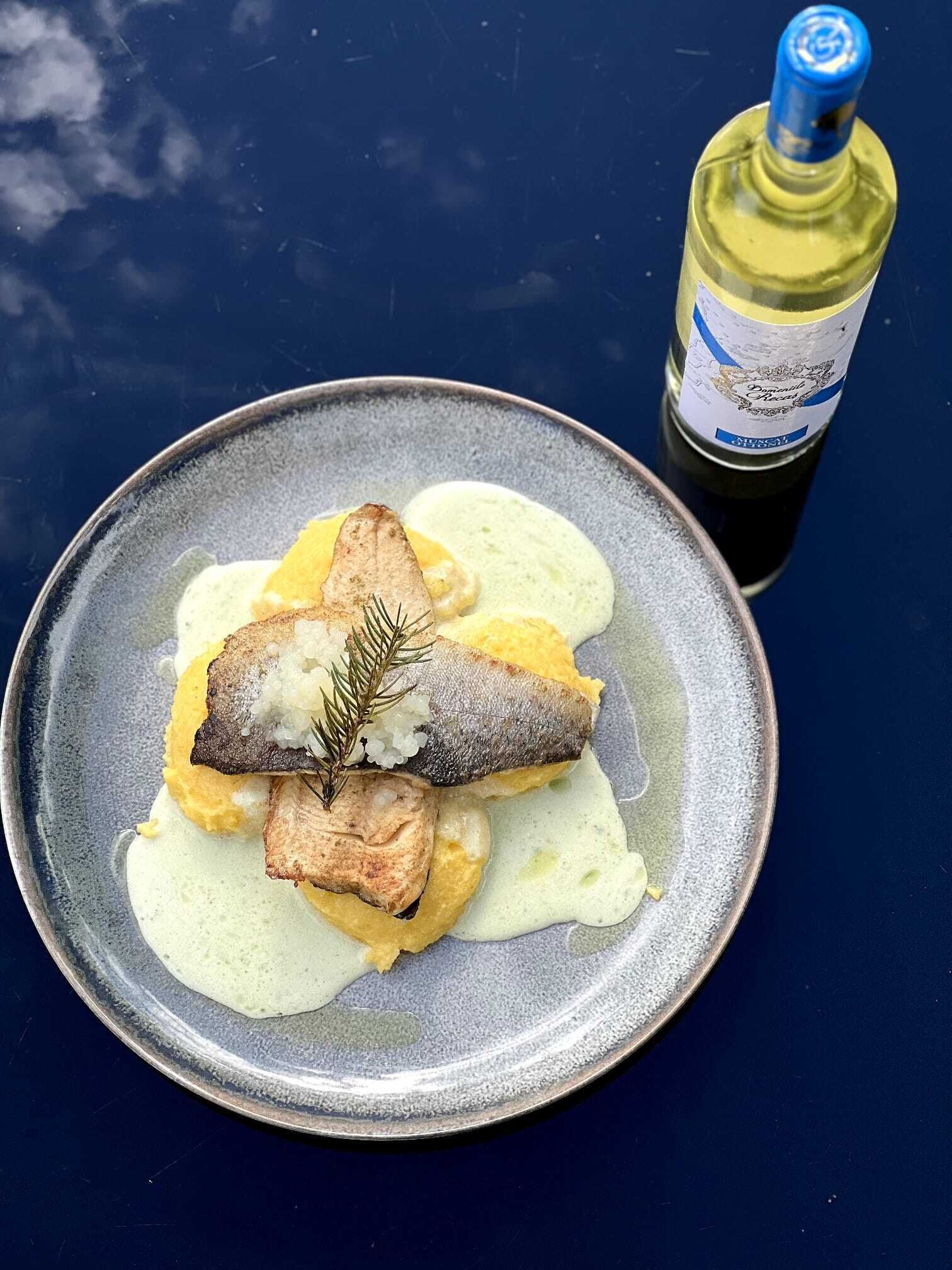 Un plat amb peix i puré de patates, un ramet de romaní i una ampolla de vi sobre una taula blava.