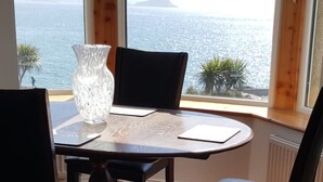 Una mesa redonda con un florero transparente, una silla negra y vista al mar a través de amplias ventanas.