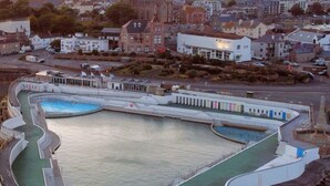 Eine Vogelperspektive auf eine Küstenstadt mit einem großen, rechteckigen Schwimmbecken, das von einer weißen Promenade umgeben ist.