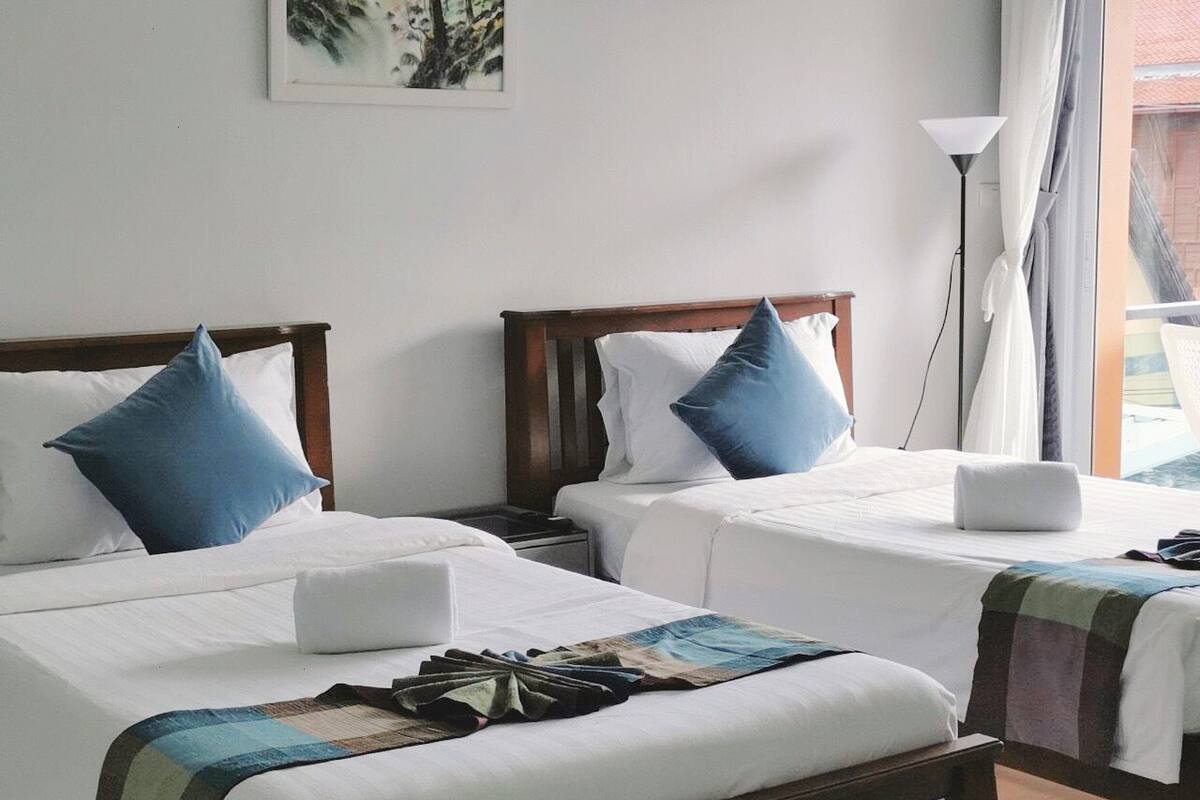 Une chambre d’hôtel avec deux lits simples, chacun doté d’une tête de lit en bois et de linge de lit blanc. Un banc se trouve au pied des lits, et une lampe de sol est visible dans un coin de la pièce.