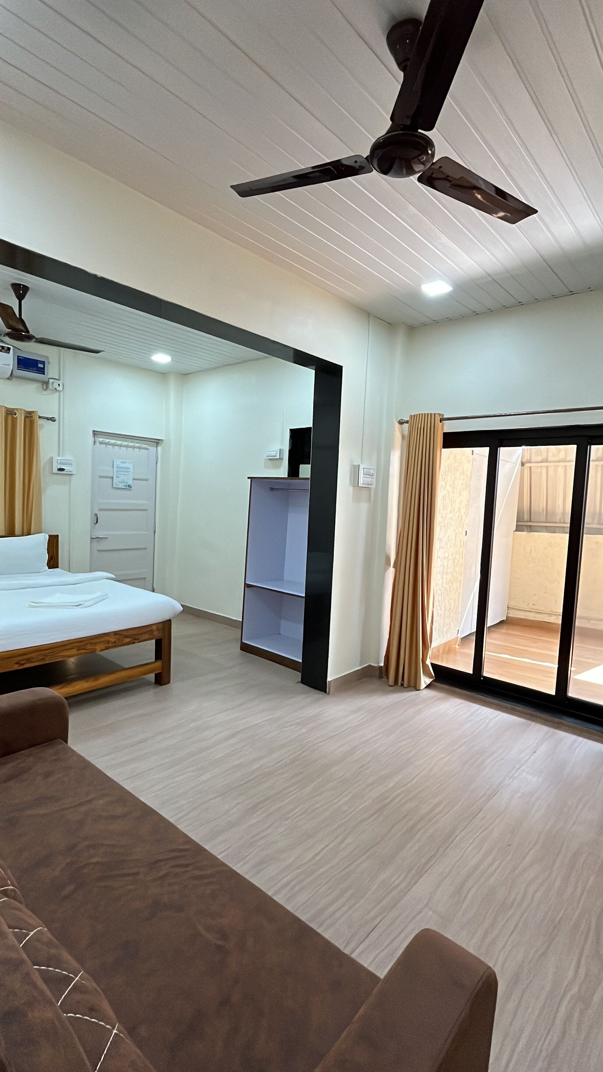 Une chambre d’hôtel équipée d’un lit, d’un bureau, d’un ventilateur de plafond, d’une armoire et d’une porte-fenêtre coulissante donnant sur l’extérieur.