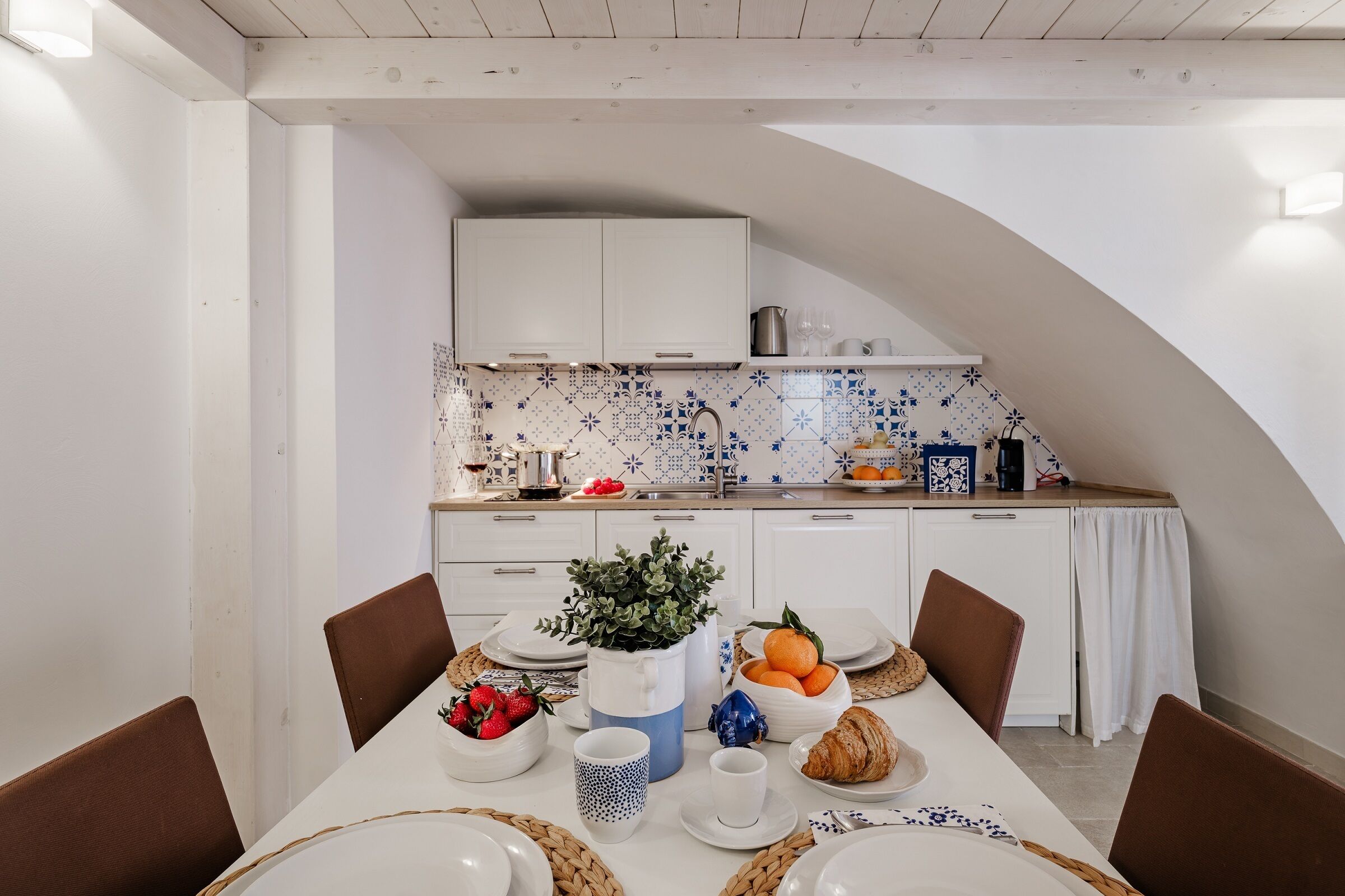 Un coin repas avec une table dressée pour deux personnes, une cuisine en arrière-plan et un mur recouvert de carreaux blancs.