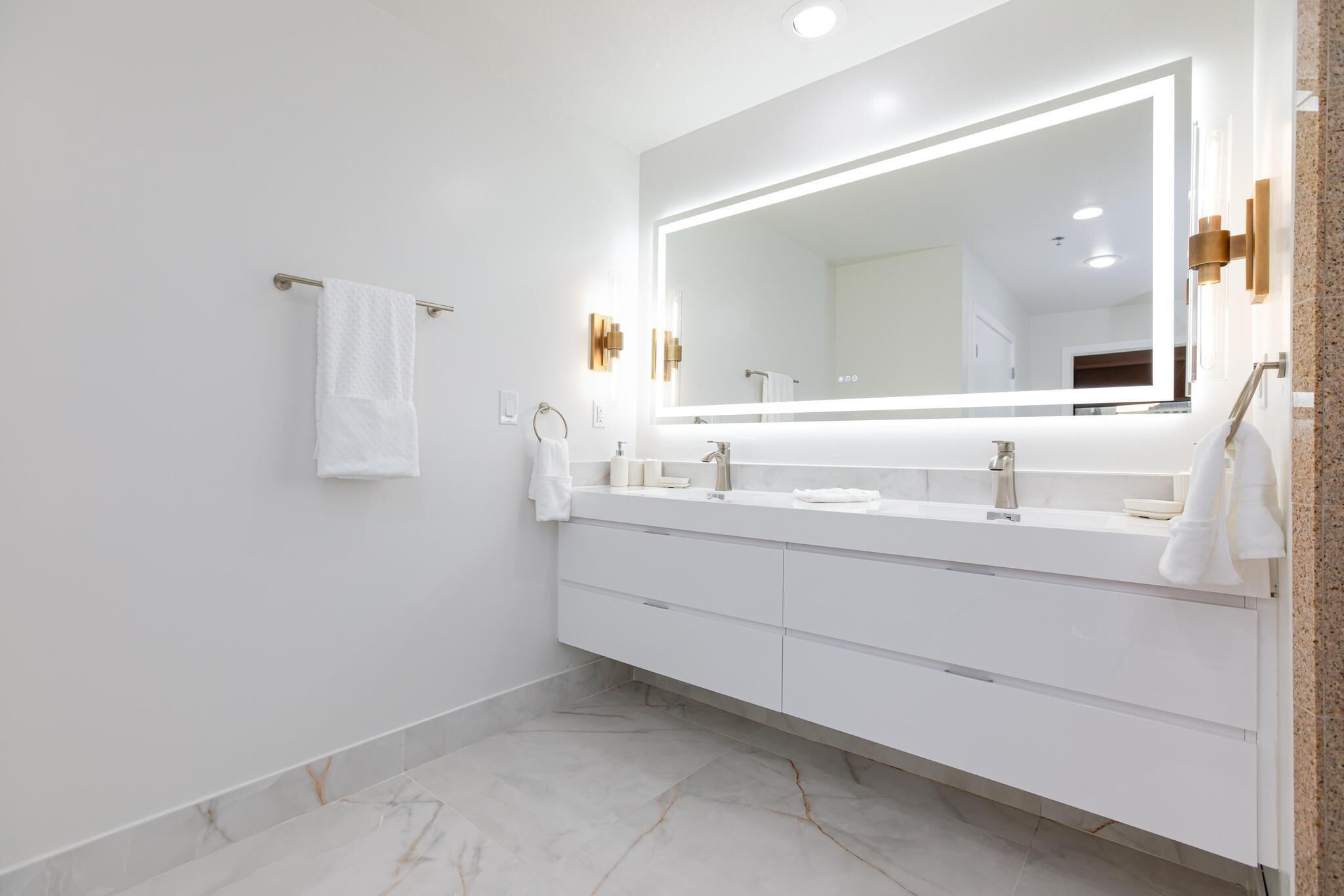 Un baño moderno con un espejo grande, tocador blanco y suelo de mármol.
