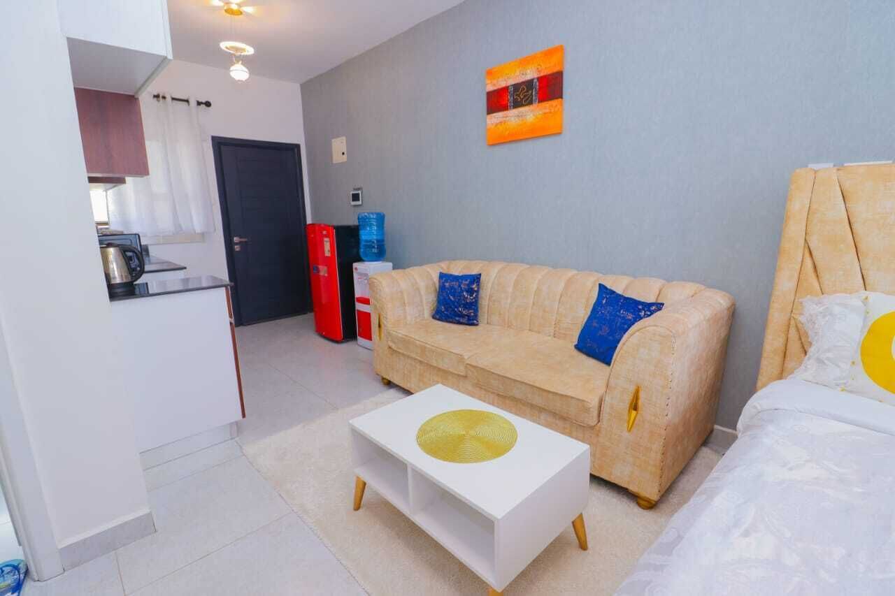 A compact living space with a kitchenette, a beige sofa with blue cushions, a white coffee table, and a bed with a yellow pillow.