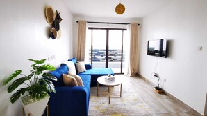 A modern living room with a blue sofa, a small round table, a potted plant, and a TV mounted on the wall. - Lux Suites Enkang Apartments Ngong Road (Nairobi)
