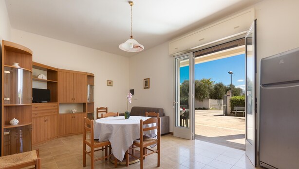 A spacious kitchen with wooden cabinets, a dining table, chairs, and a large sliding glass door leading to an outdoor area. - 3847 Villetta Le Largure B by Barbarhouse (Melendugno)