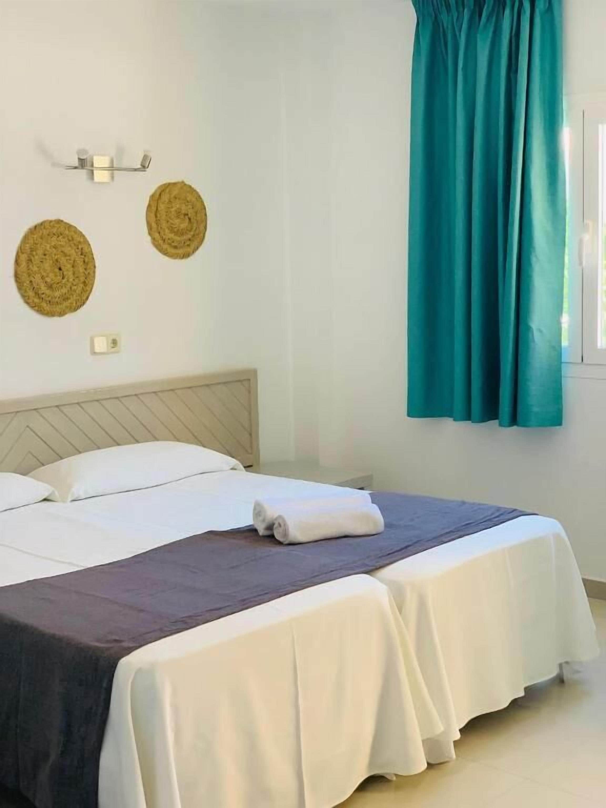 Una cama bien hecha con sábanas blancas y una colcha oscura, una cortina color turquesa y dos adornos de pared tejidos.