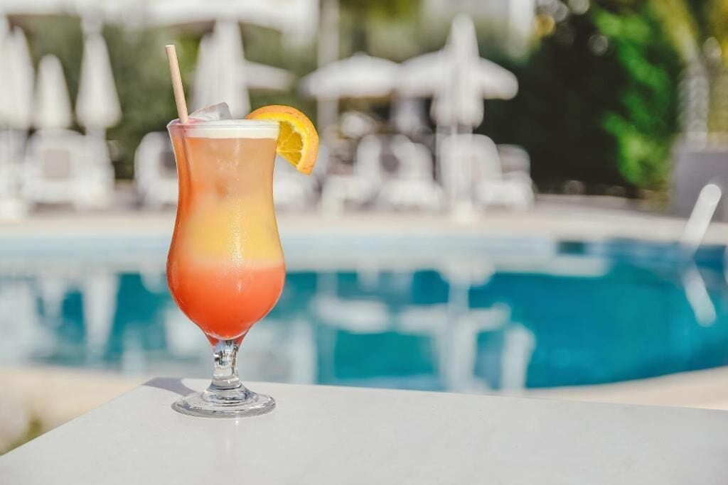 Un cocktail in un bicchiere con una cannuccia e una fetta d'arancia, posato su un tavolo vicino a una piscina con lettini e ombrelloni sullo sfondo.